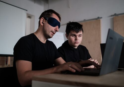 Participant cu ochelari opaci este îndrumat de un voluntar cu deficiențe de vedere în utilizarea laptop-ului.