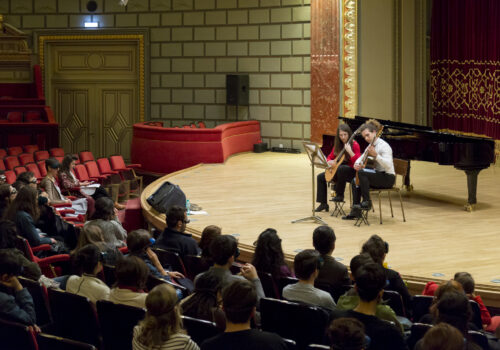 Cadrul cu sala și publicul și doi interpreți pe scenă la chitară