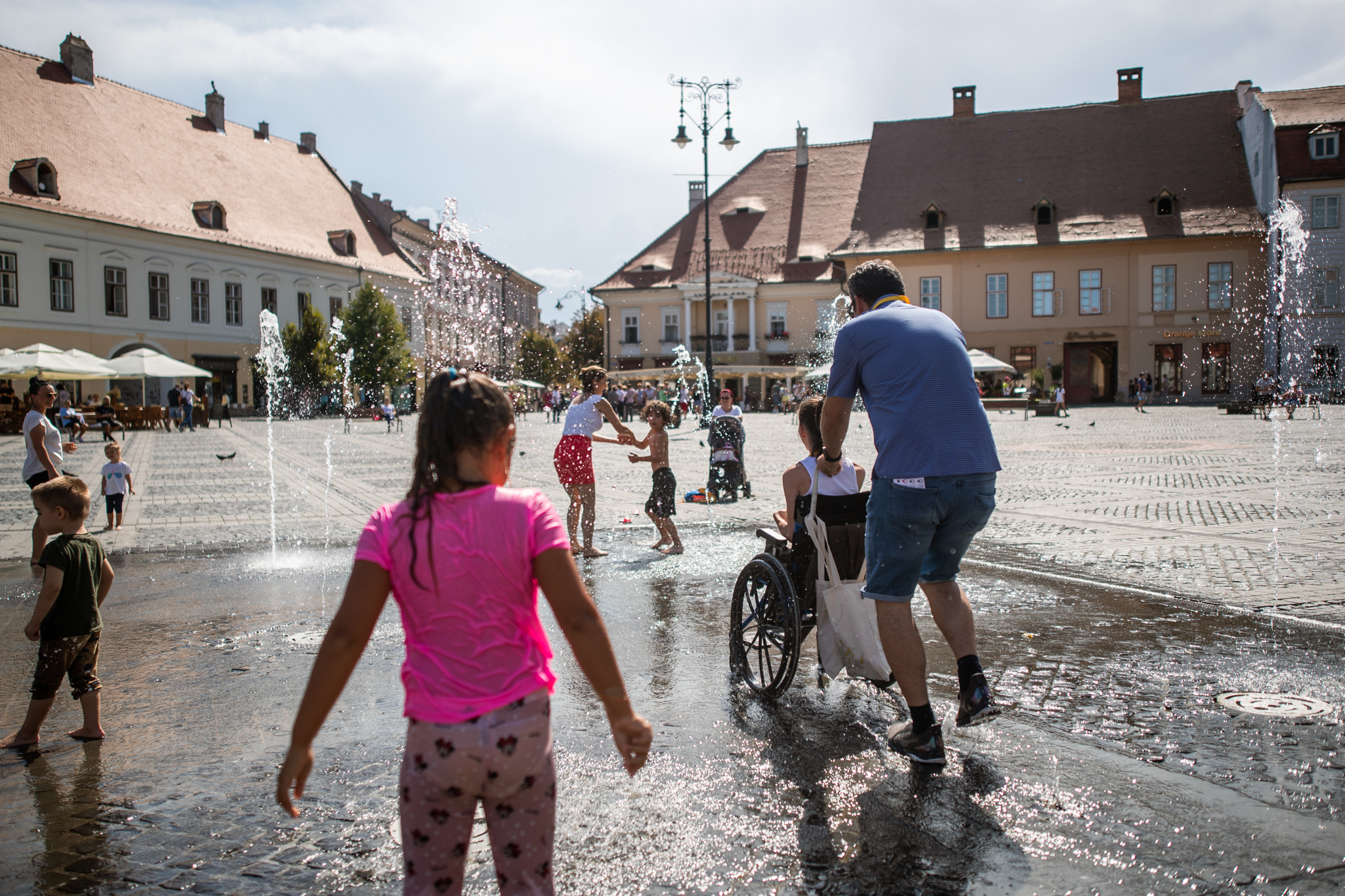 Eugen împinge scaunul cu rotile al lui Teo printre picăturile de apă din Piața Mare din Sibiu