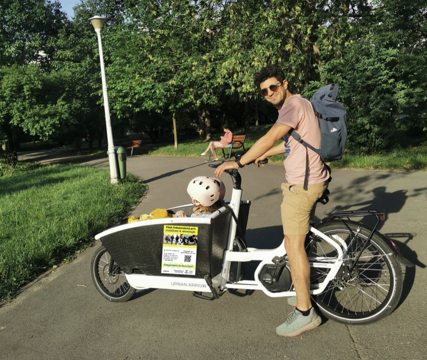 Alex Călin surprins în parc pe o bicicletă cargo în care își plimbă copilul care poartă o cască de protecție roz.