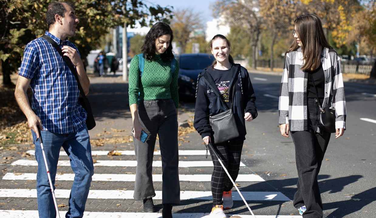 Four persons are crossing the street. Two of them, a boy and a girl, are visually impaired and use a white cane