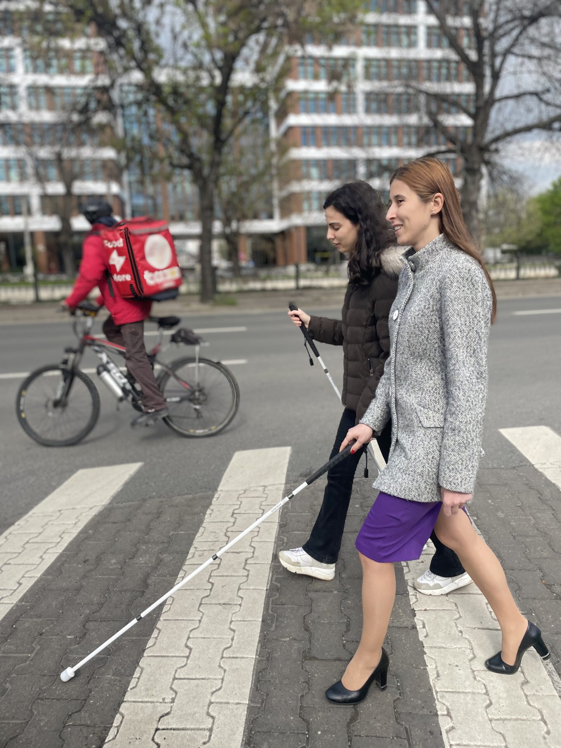 Two girls are crossing the street. The photo is taken from the profile. One of the girls is using the white cane. In the baclground we can see blocks and a delivery person riding a bike. 
