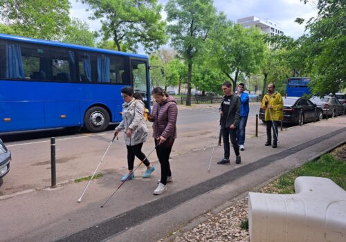 Patru tineri merg cu bastonul alb pe trotuar. Pe carosabil, un autobuz mare și albastru.