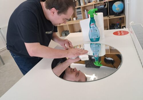 Un tânăr șterge o oglindă rotundă așezată pe o masă. Zâmbește. Are lângă el șervețele și un spray de geamuri.
