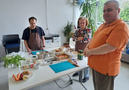 Trei persoane cu dizabilități de vedere stau în picioare în jurul unei mese pe care sunt așezate tocătoare și castroane cu ingrediente pentru supă înainte de un atelier de gătit.