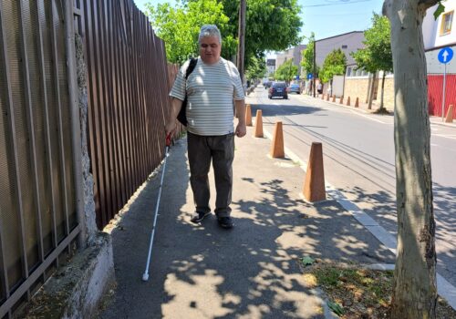 Un bărbat merge singur pe trotuar cu bastonul, ținând reper un gard. Afară e soare.