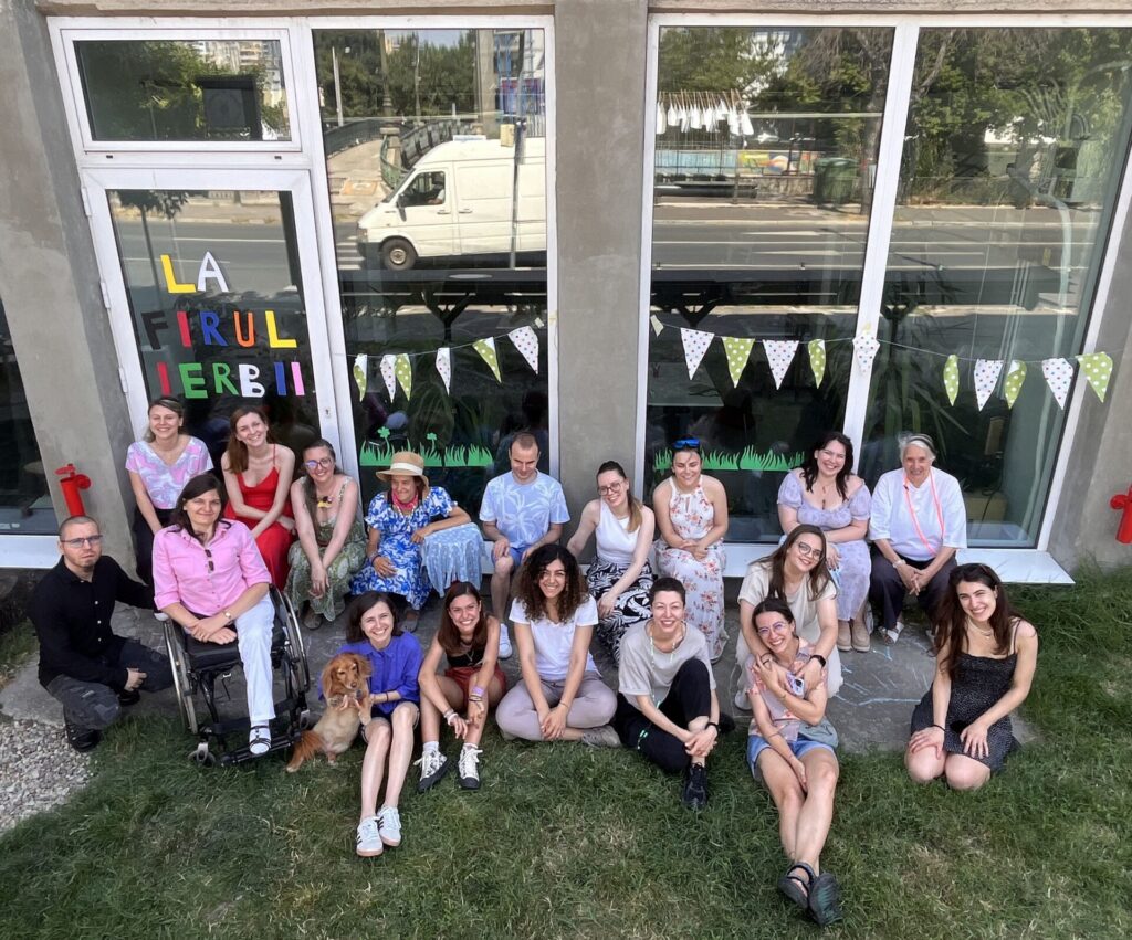 Group photo in the Grassroots Garden, after an inclusive creative workshop. People with and without disabilities alongside members of the AMAIS and Jos Boys teams, a British architect, and a little dog. They are smiling. Behind them is the building with tall windows decorated with small flags and other ornaments.