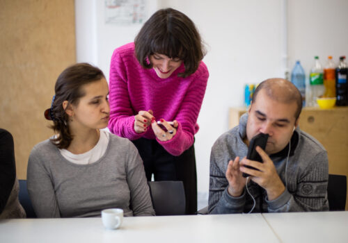 Două persoane cu dizabilități de vedere stau la masă cu câte un telefon și o tânără fără dizabilități de vedere stă în spatele lor și vorbește cu ei.