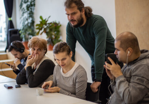 Fotografie din timpul unui focus grup. Patru persoane cu dizabilități de vedere stau la masă cu telefoanele în mâna și căștile în urechi. În spatele lor, în picioare, stă Alex cucu.