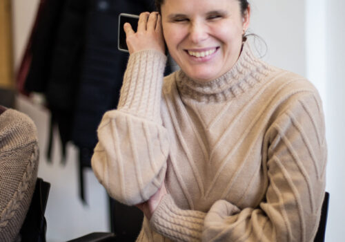 Loredana, persoană cu dizabilități de vedere, poartă pulover și are telefonul la ureche. Ascultă ce citește sinteza vocală.