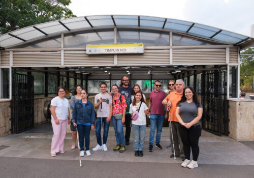 Trainerii de la training of trainers, invitații, trainerii Ana Costache și Alex Matei în fața stației de metrou Timpuri Noi. Toți zâmbesc.