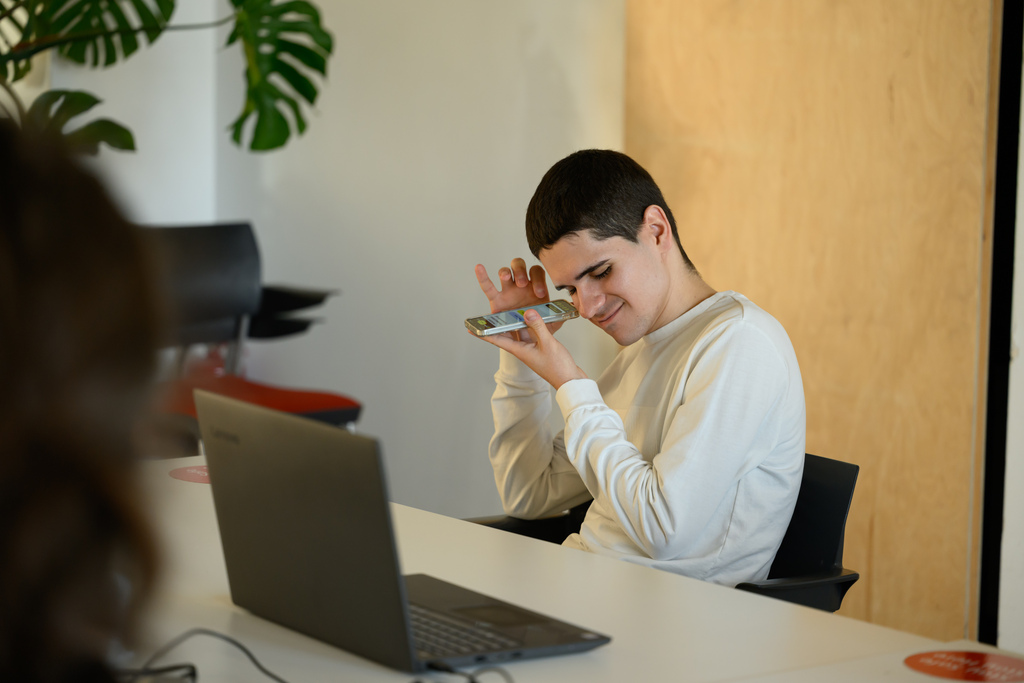 alex matei stă la o masă în fața unui laptop și are telefonul ridicat la ureche, să asculte cititorul de ecran. zâmbește.