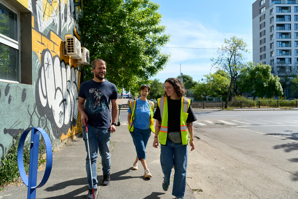 De la stânga la dreapta, alex cucu cu baston alb, nancy și ana costache, trainer de mobilitate merg pe trotuar. e soare și frumos.